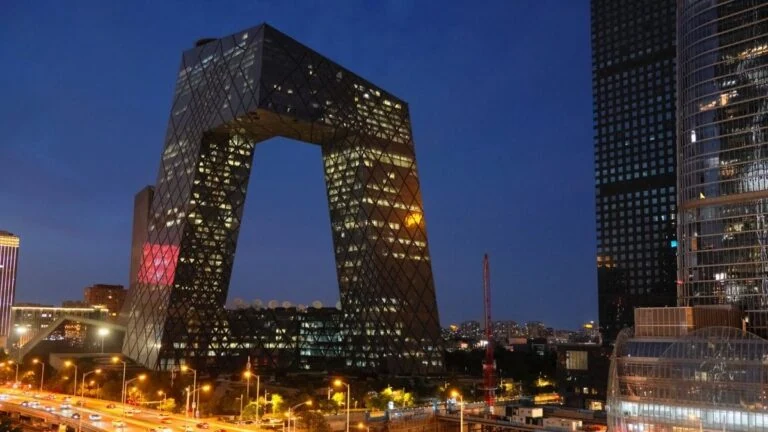 Nighttime cityscape showing the Beijing CCTV Headquarters with illuminated windows and surrounding buildings along a busy street.