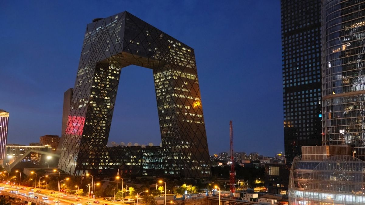 Nighttime cityscape showing the Beijing CCTV Headquarters with illuminated windows and surrounding buildings along a busy street.