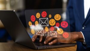 Person typing on a laptop with envelope icons and orange/red angry emojis hovering above, illustrating hostile emails.