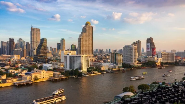 Skyline of a riverfront city with tall buildings and boats on the river at sunset.