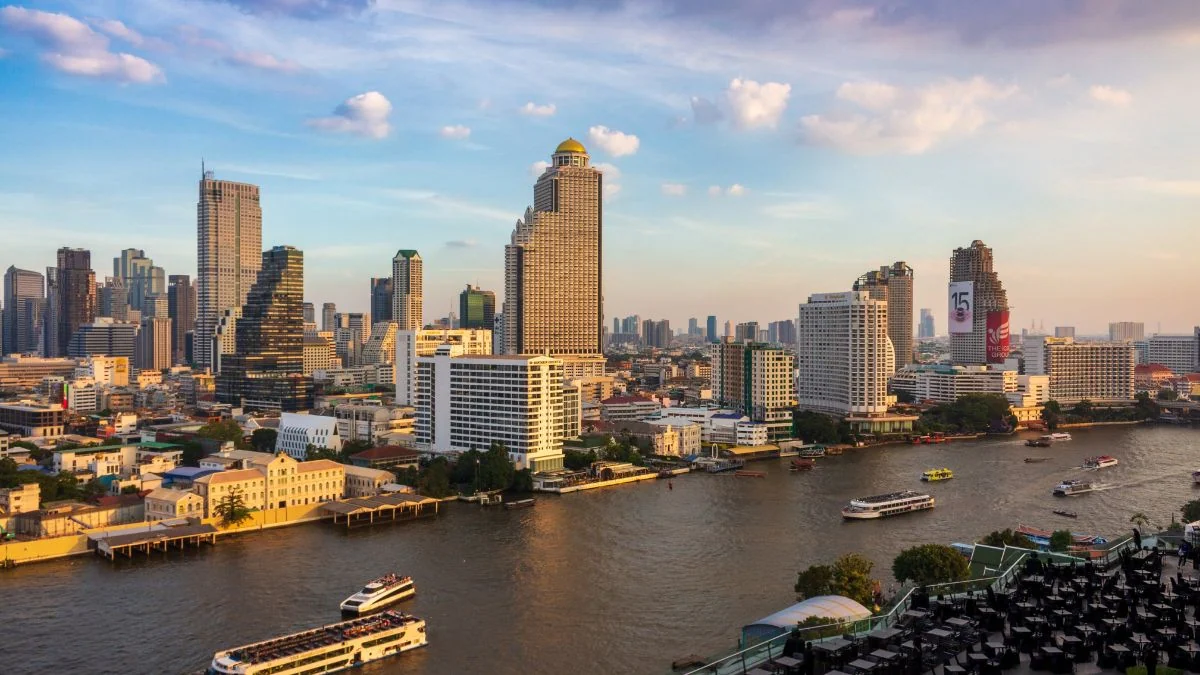 Skyline of a riverfront city with tall buildings and boats on the river at sunset.