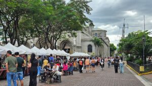 Outdoor market with white vendor tents along a cobbled street in front of a stone church, people shopping and strolling.
