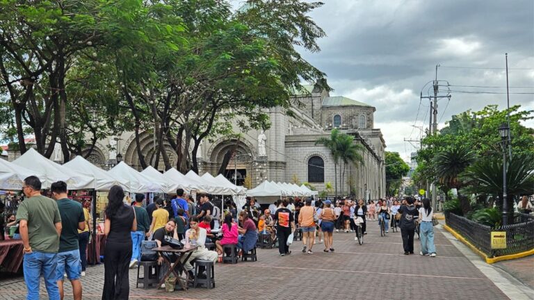 Outdoor market with white vendor tents along a cobbled street in front of a stone church, people shopping and strolling.