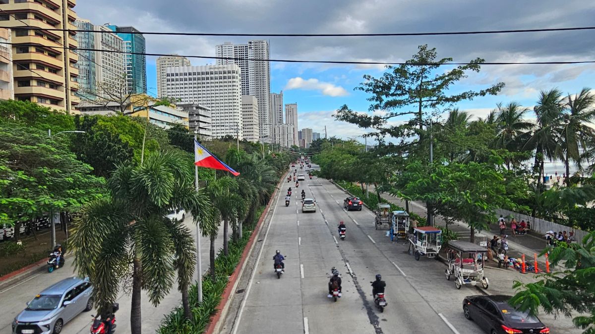 Urban boulevard with palm trees and a Philippine flag along a busy multilane road in a city setting with high-rise buildings in the distance.