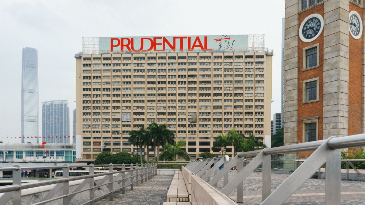 Large beige office building with a red PRUDENTIAL sign on top, palm trees in front, and a clock-tower to the right; foreground walkway with railings.