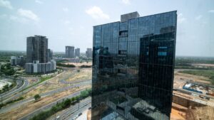 Tall glass office tower dominates the foreground, its reflective façade mirroring nearby buildings and a road network below on a sunny day.