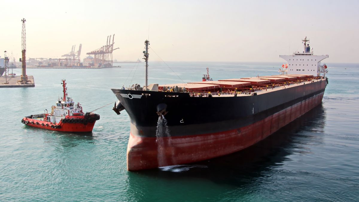 Large black-and-red bulk carrier being guided by a small tugboat at a port, with cranes in the distance and calm turquoise water.