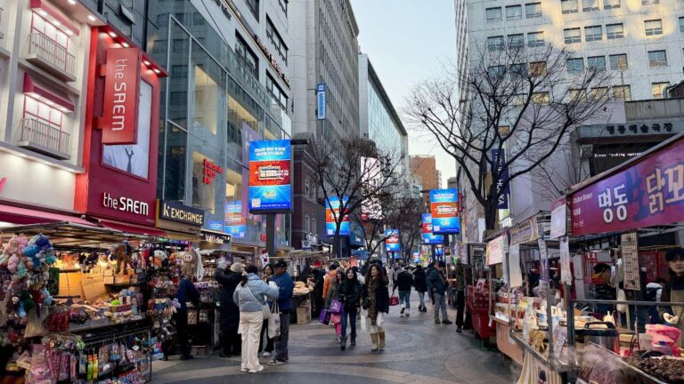 Bustling street market in an urban shopping district with stalls selling toys and snacks and shoppers browsing.