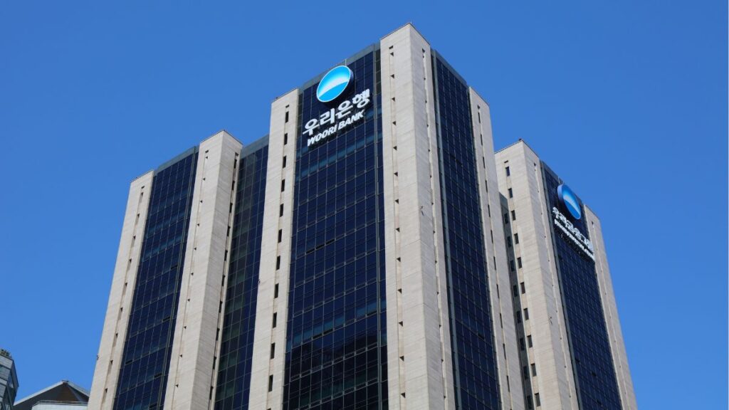 Modern high-rise bank building featuring Woori Bank logos on a blue glass facade against a clear blue sky.