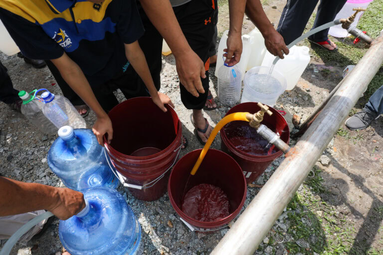 People fill large water containers from a hose at an outdoor work site on rocky ground.