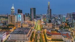 Aerial view of a city at blue hour with a wide, tree-lined boulevard and surrounding tall office towers and illuminated streets.