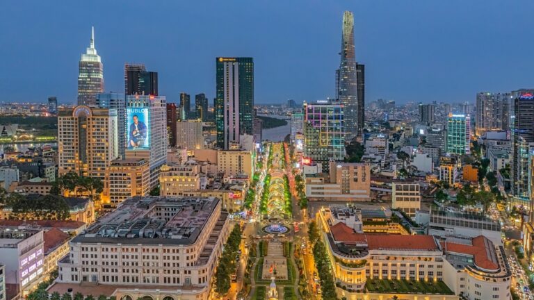 Aerial view of a city at blue hour with a wide, tree-lined boulevard and surrounding tall office towers and illuminated streets.