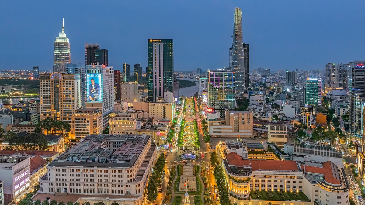 Aerial view of a city at blue hour with a wide, tree-lined boulevard and surrounding tall office towers and illuminated streets.
