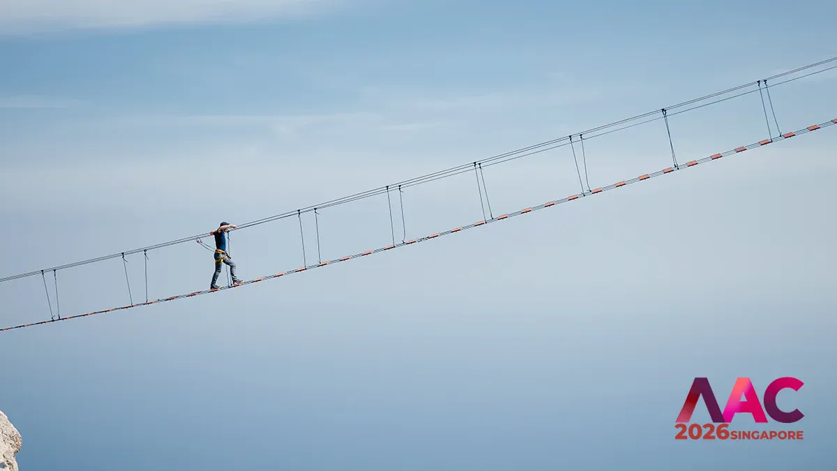 A person wearing a harness walks along a long suspended rope bridge high above, with a clear blue sky and the AAC 2026 Singapore branding in the bottom-right corner.