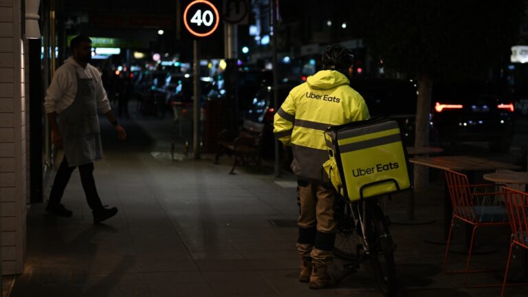 Uber Eats courier in a bright yellow reflective jacket with a delivery bag on a bicycle at night on a city street.