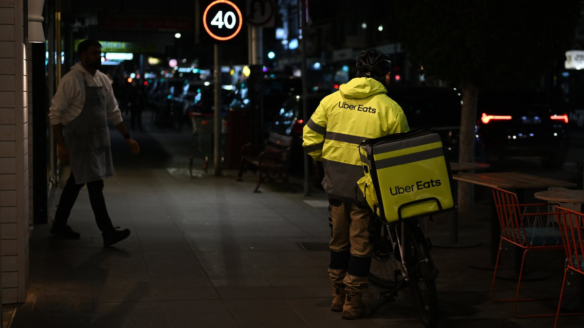 Uber Eats courier in a bright yellow reflective jacket with a delivery bag on a bicycle at night on a city street.