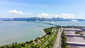 Aerial view of a long bridge crossing a calm bay toward distant mountains, with a green parkway and multi-lane road below.