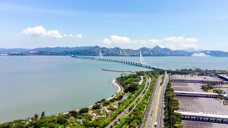 Aerial view of a long bridge crossing a calm bay toward distant mountains, with a green parkway and multi-lane road below.
