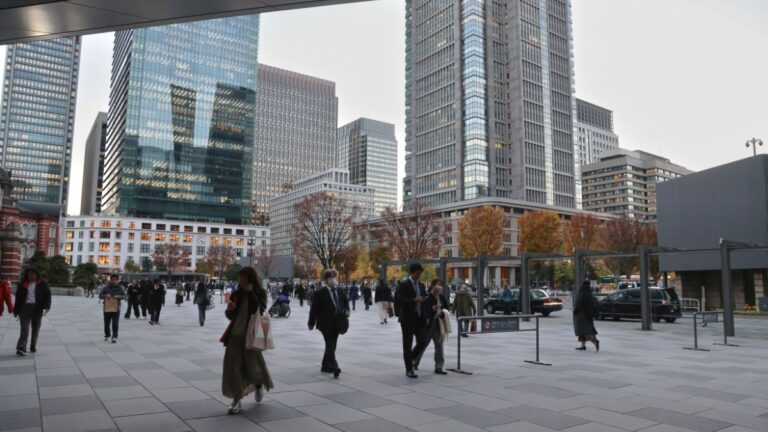 Busy urban plaza with pedestrians and modern glass skyscrapers in the background during autumn sunlightfall colors in trees behind them