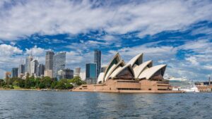 Iconic white sails of the Sydney Opera House beside Sydney Harbour with a city skyline and blue sky behind it.