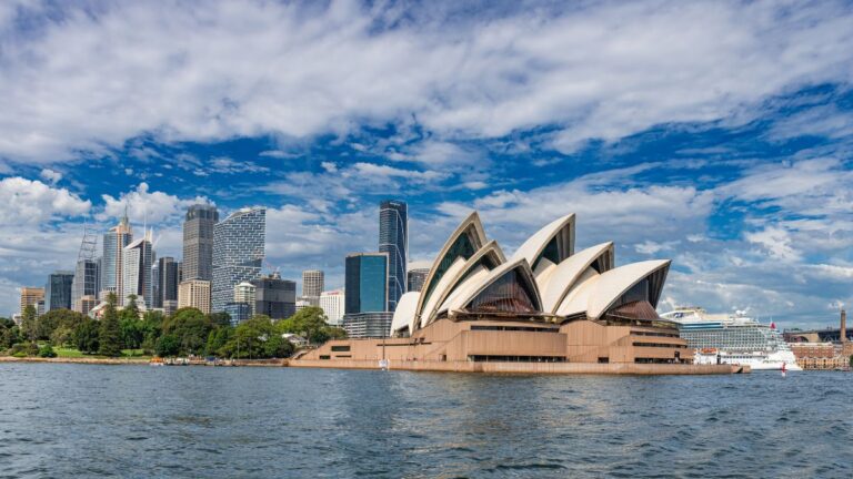 Iconic white sails of the Sydney Opera House beside Sydney Harbour with a city skyline and blue sky behind it.