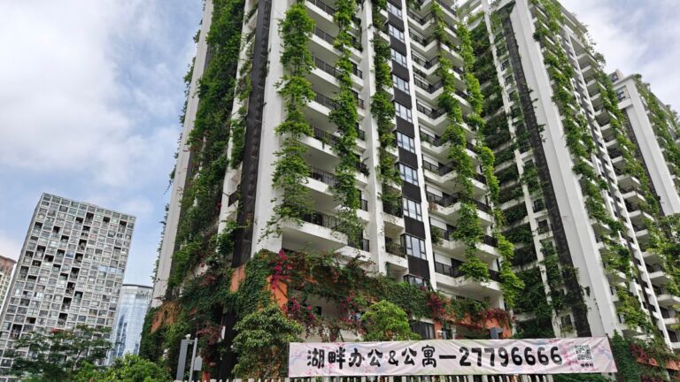 Tall modern high-rise apartment building covered in vertical greenery along balconies, with a large banner at the ground level.