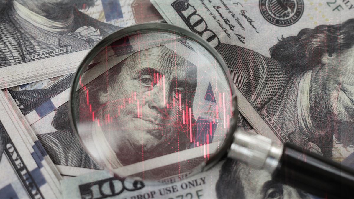Magnifying glass over US dollar bills with red stock chart lines, suggesting financial analysis or scrutiny.