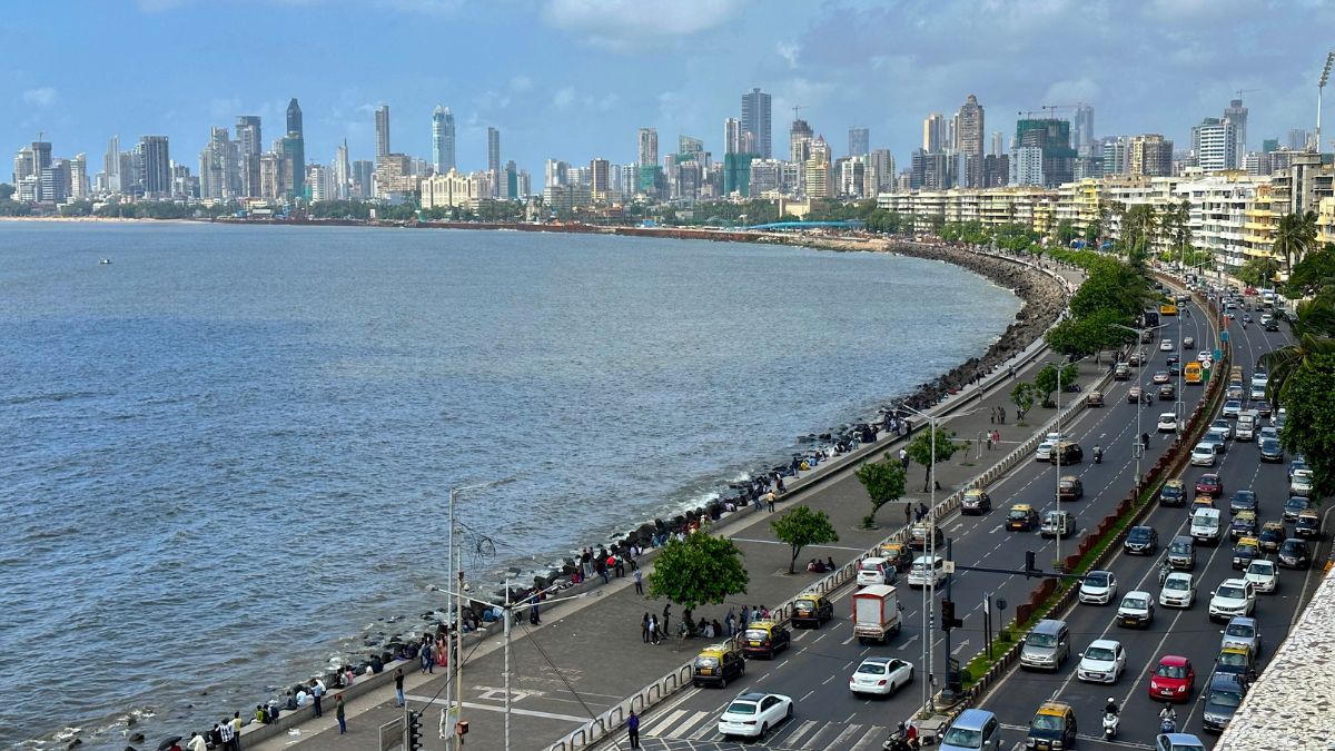 Seaside cityscape with a curved waterfront promenade, people walking along the railing, and a busy coastal road lined with cars; skyline in the distance.
