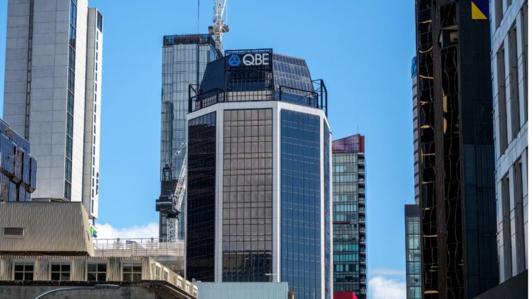 Modern city skyline featuring glass office towers under a clear blue sky, with a prominent QBE sign on the central building.