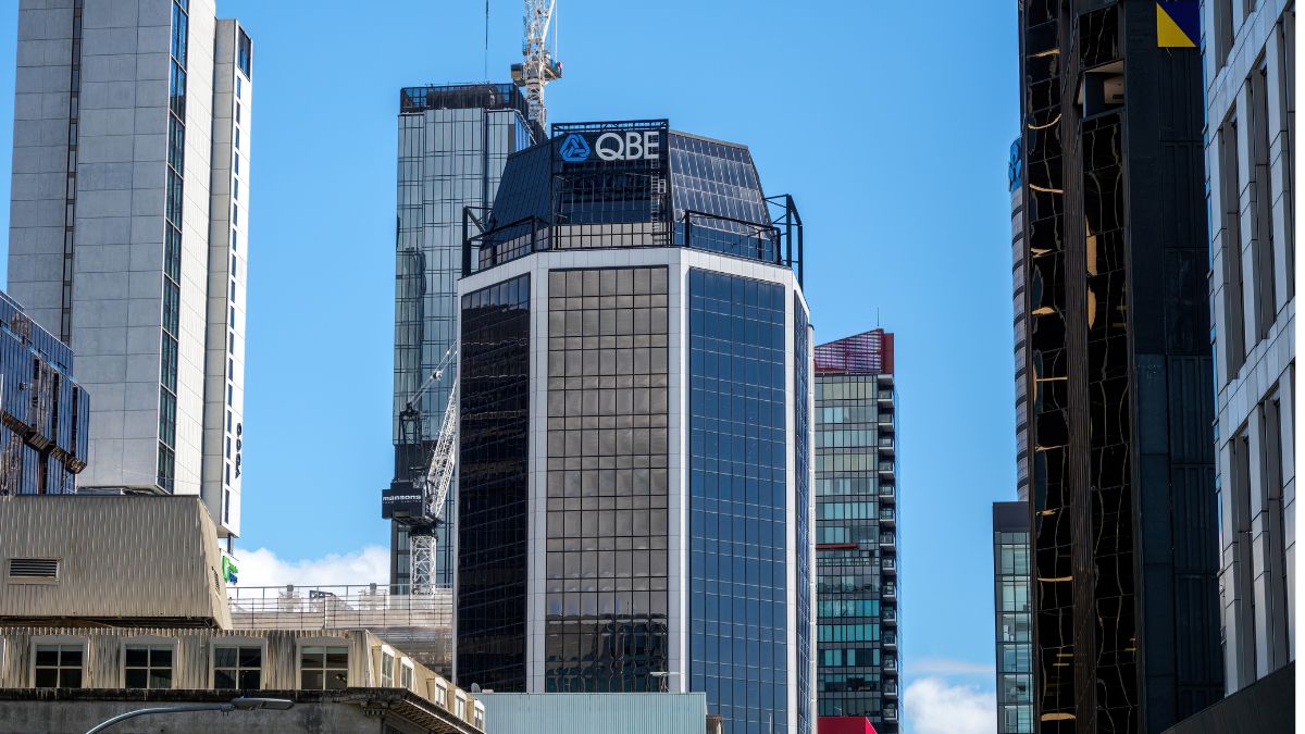 Modern city skyline featuring glass office towers under a clear blue sky, with a prominent QBE sign on the central building.
