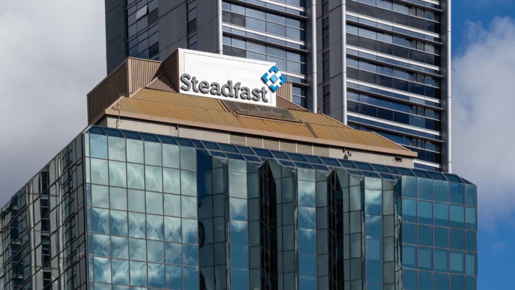 Glass-front office building with a prominent Steadfast sign on the roof and smokey-brown roofing, reflecting a blue sky in the glass panels