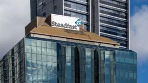 Glass-front office building with a prominent Steadfast sign on the roof and smokey-brown roofing, reflecting a blue sky in the glass panels