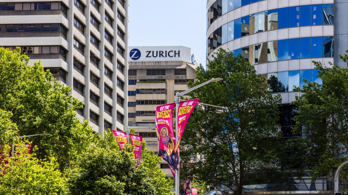 Urban street scene with tall office buildings and a Zurich sign atop a beige tower; pink banners hang from street lamps.