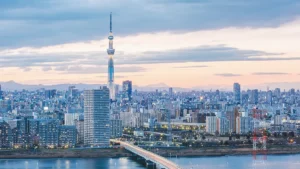 Dusk cityscape of Tokyo with the tall Tokyo Skytree rising above a dense cluster of buildings and a river in the foreground.