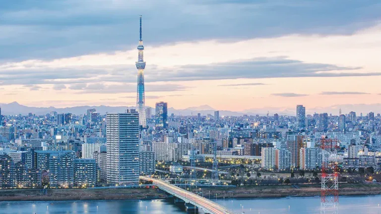 Dusk cityscape of Tokyo with the tall Tokyo Skytree rising above a dense cluster of buildings and a river in the foreground.