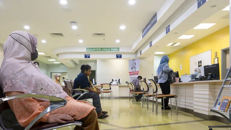 Waiting area in a clinic with people seated and a staff member at a bright yellow reception desk under a 'KAUNTER JANJI TEMU' sign