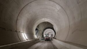Interior of a large circular tunnel under construction; a small excavator sits on a dirt floor with concentric tunnel rings receding into the distance.
