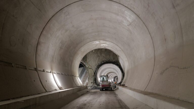 Interior of a large circular tunnel under construction; a small excavator sits on a dirt floor with concentric tunnel rings receding into the distance.