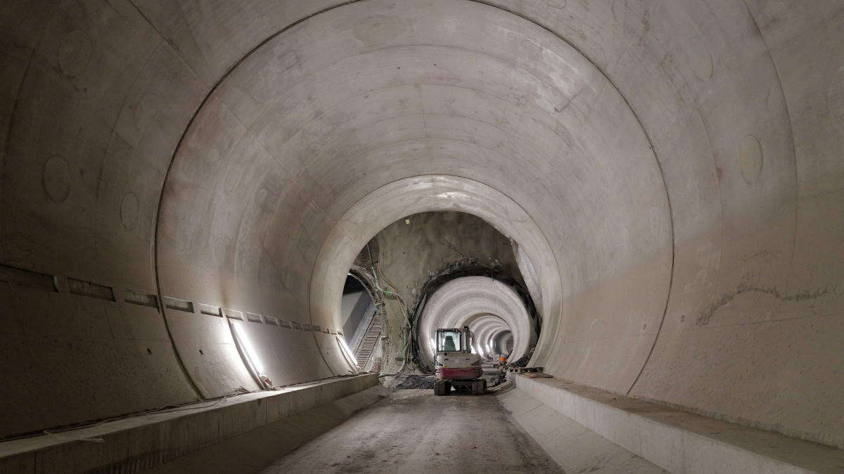 Interior of a large circular tunnel under construction; a small excavator sits on a dirt floor with concentric tunnel rings receding into the distance.