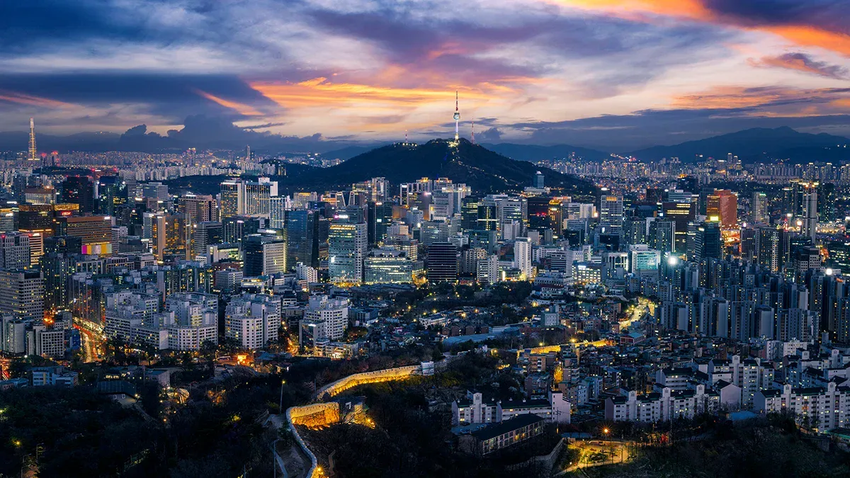 Wide city skyline at dusk with a hill and a tall tower on top, colorful sunset in the clouds above Seoul-like cityscape.
