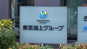 Company sign for Tokio Marine Group with blue and yellow logo, displaying Japanese text and the English group name underneath, set among plants outside a building.