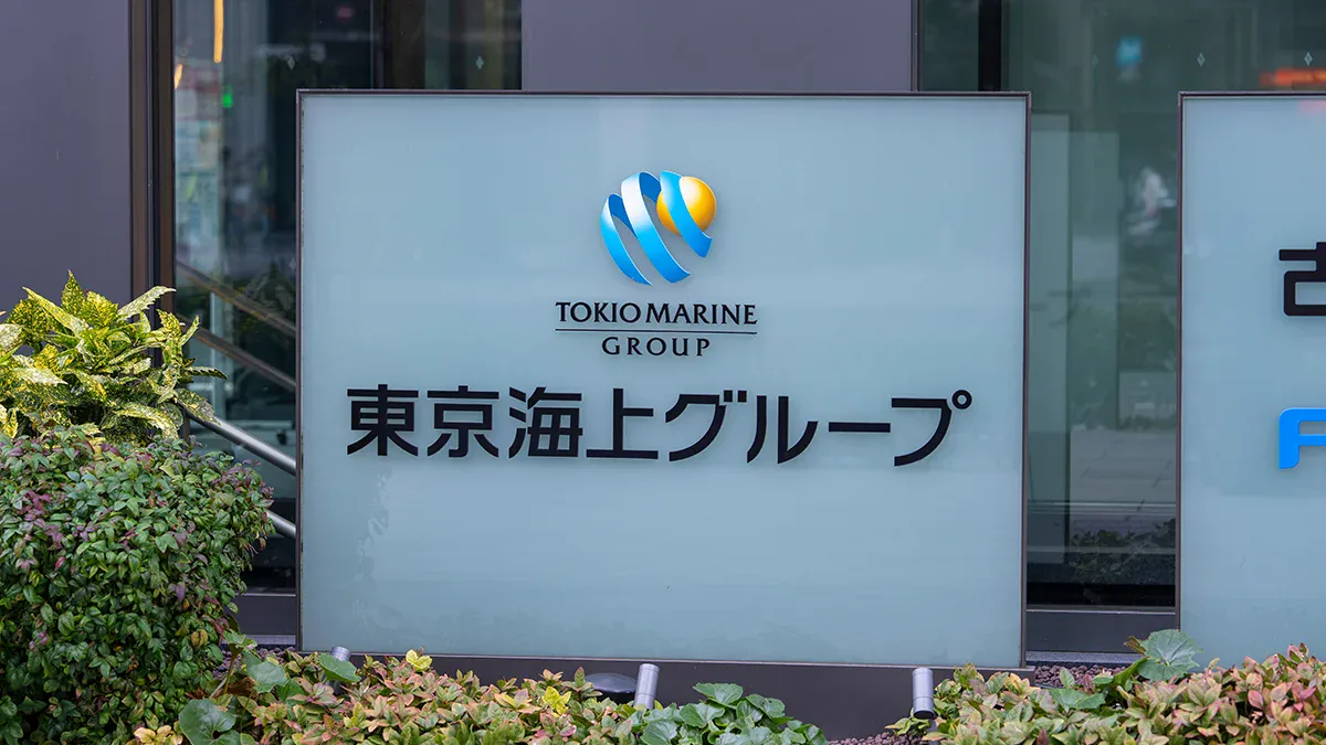 Company sign for Tokio Marine Group with blue and yellow logo, displaying Japanese text and the English group name underneath, set among plants outside a building.
