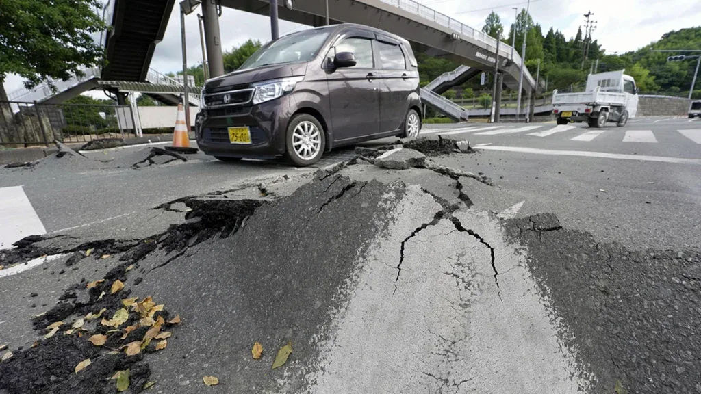 A damaged road with a large cracked slump hole; a dark minivan parked nearby under an overpass, with traffic cones around the area.