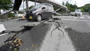 A damaged road with a large cracked slump hole; a dark minivan parked nearby under an overpass, with traffic cones around the area.