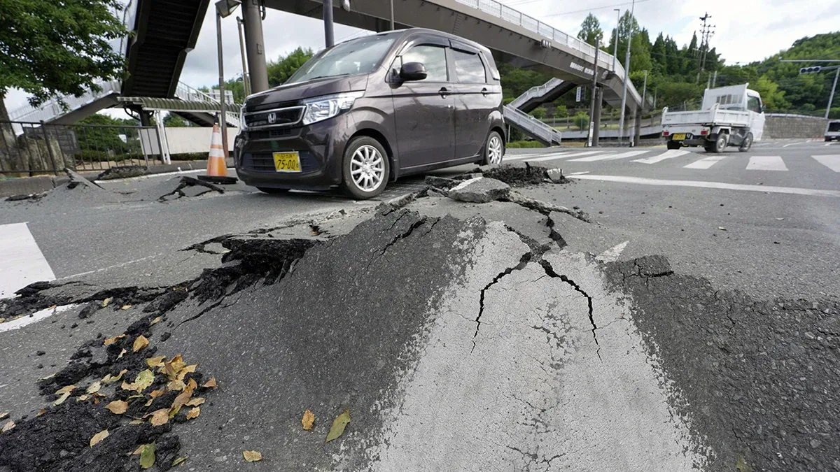 A damaged road with a large cracked slump hole; a dark minivan parked nearby under an overpass, with traffic cones around the area.