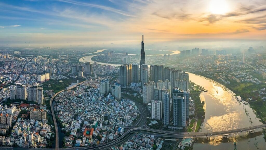 Aerial view of a densely built city with a winding river, high-rise towers, and a sunset sky above the skyline.