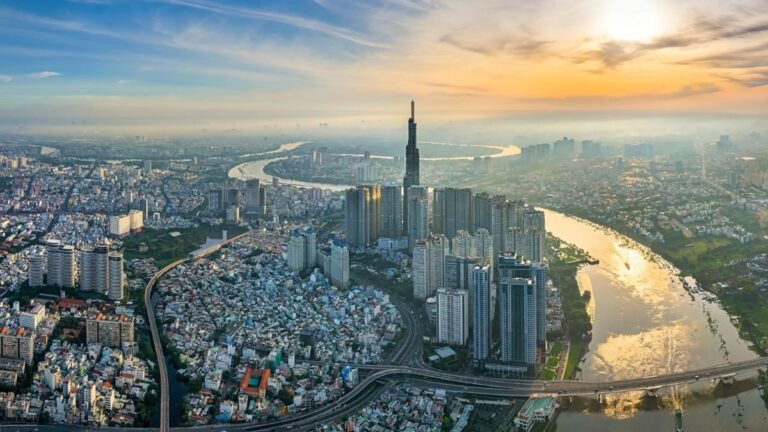 Aerial view of a densely built city with a winding river, high-rise towers, and a sunset sky above the skyline.