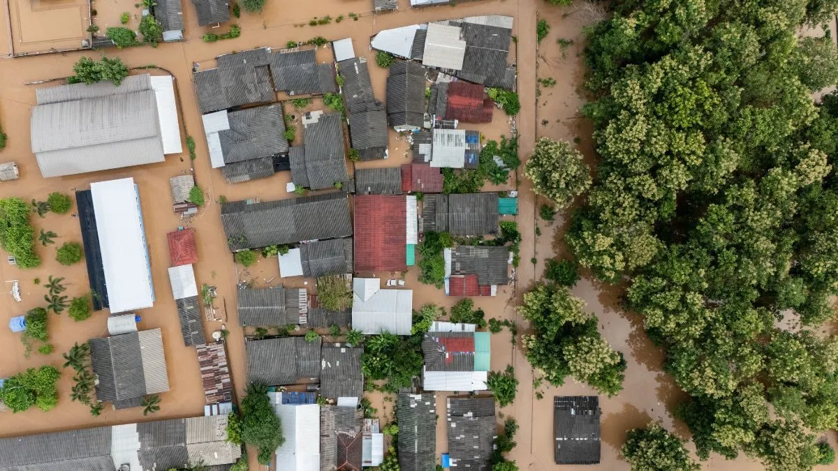 Aerial view of muddy floodwaters surrounding homes, roads and trees in a densely built settlement.