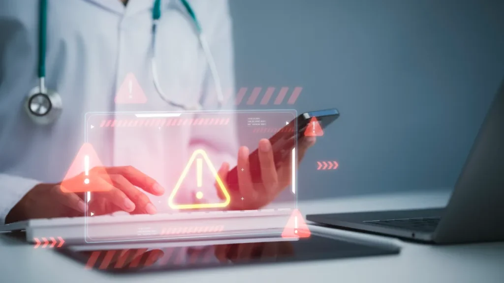Healthcare professional using a tablet beside a laptop, with digital warning symbols suggesting a data security breach.
