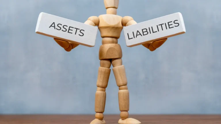 Wooden mannequin holds signs labelled “ASSETS” and “LIABILITIES”.